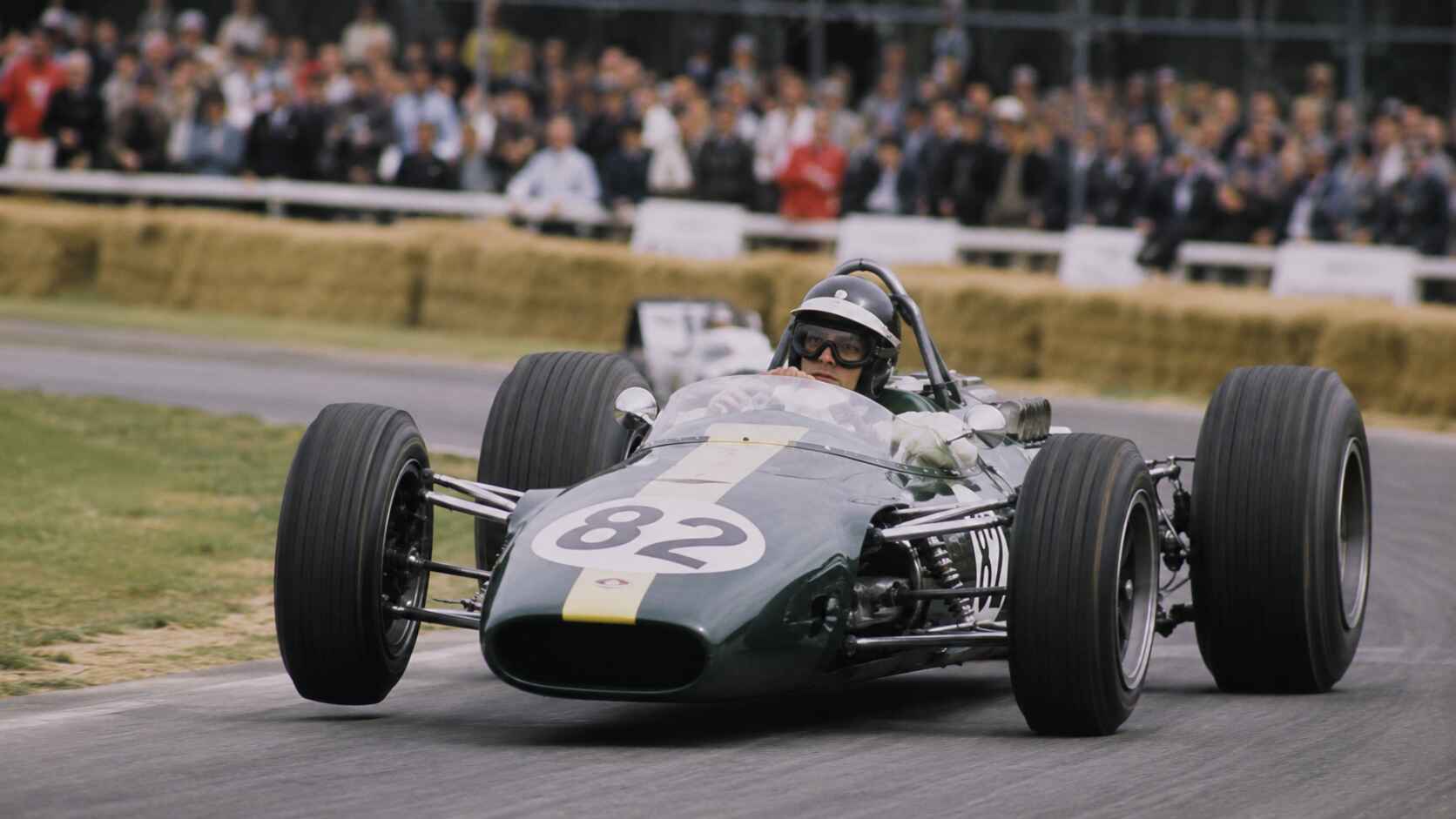 Voiture de course monoplace d'époque vert foncé avec le numéro 82 et bande jaune, en action sur une piste asphaltée sinueuse.